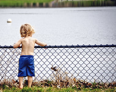 boy leaning on gray metal fence beside lake