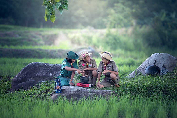 two boy scout and one girl scout cooking on pot using stove surrounded by green grass field during daytime