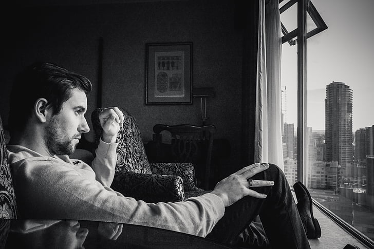 man sitting on sofa chair while staring through the window grayscale photo