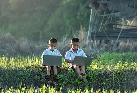 two boys in white dress shirt sitting on grass using laptop computers