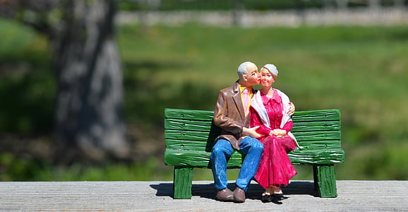 shallow focus photography of man wearing brown formal suit jacket and woman wearing red long-sleeved dress sitting on green bench ceramic figurine