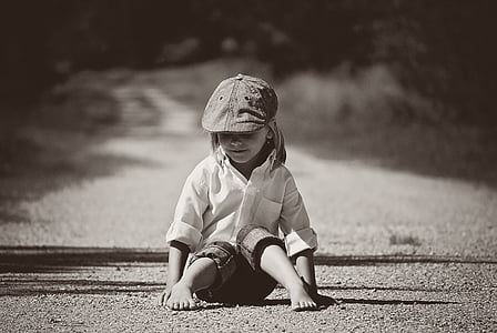 greyscale photography of boy wearing dress shirt and newsboy cap sitting on ground at daytime