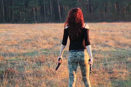 woman wearing black cold-shoulder quarter-sleeved shirt with green and black camouflage pants holding two black semi-automatic pistols standing on brown and green grass field