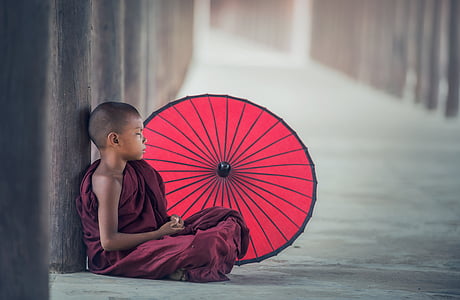 photo of child monk in maroon robe sitting on floor beside red parasol