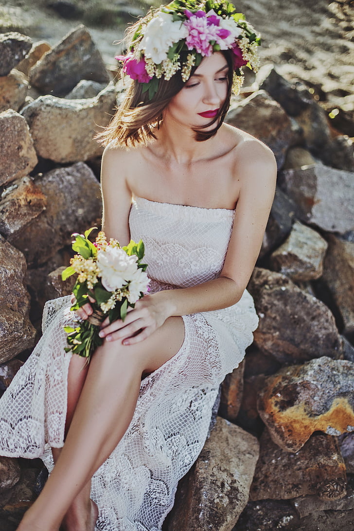 woman wearing white lace tube dress sitting on rock holding white flower bouquet
