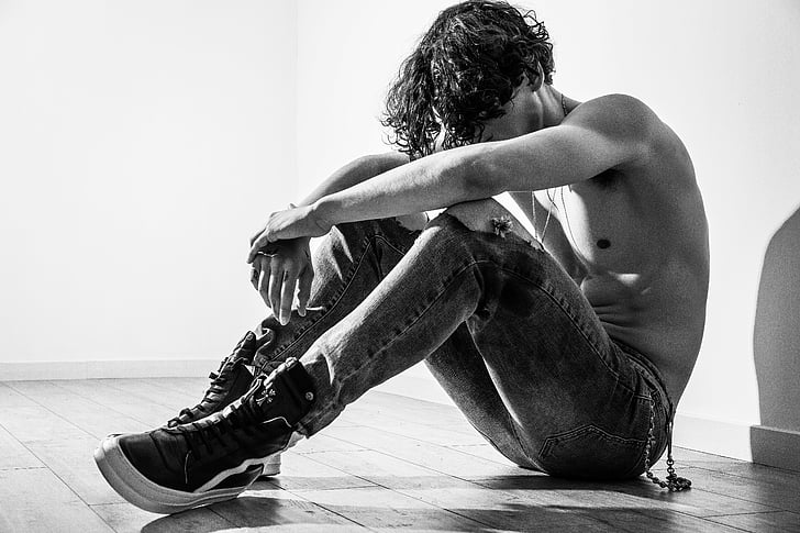 greyscale photo of topless man sitting on floor near wall inside room