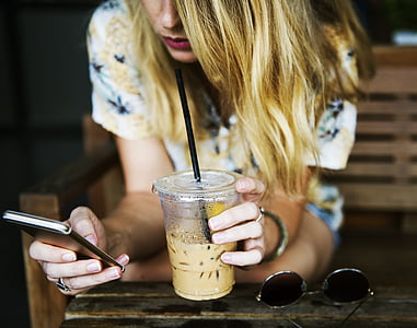 selective focus photography of blonde haired woman sipping yellow liquid while using smartphone