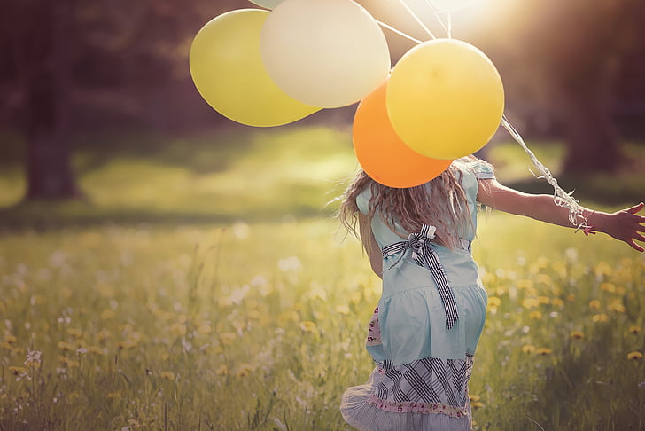 selective focus photography of girl in blue dress holding yellow balloons while playing on grass field