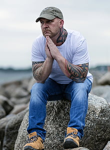 tattooed man wearing gray baseball cap, white crew-neck t-shirt, and blue denim jeans sitting on gray boulder under gray clouds