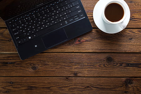 black laptop computer beside white ceramic teacup with saucer on brown wooden surface