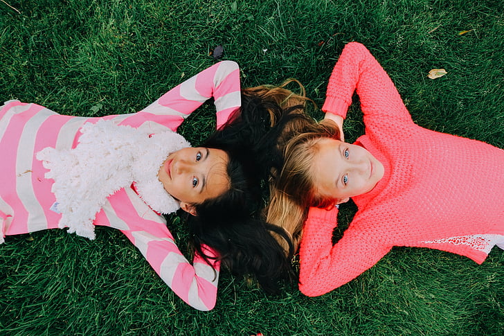 two girls wearing pink and white long-sleeved shirts lying on grass during daytime