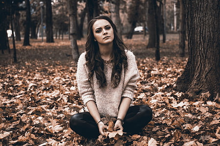 woman in gray sweater and black pants sitting on ground full of leaves