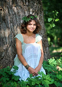 woman wearing teal scoop-neck cap-sleeved dress sitting in front of tree surrounded by green leaf plants
