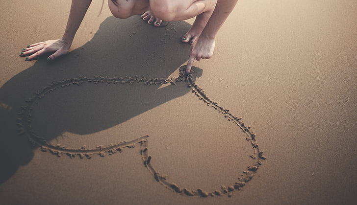 person drawing heart shape on sand