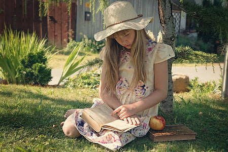 woman in yellow floral dress reading book under brown tree with brown sunhat beside red honeycrisp apple