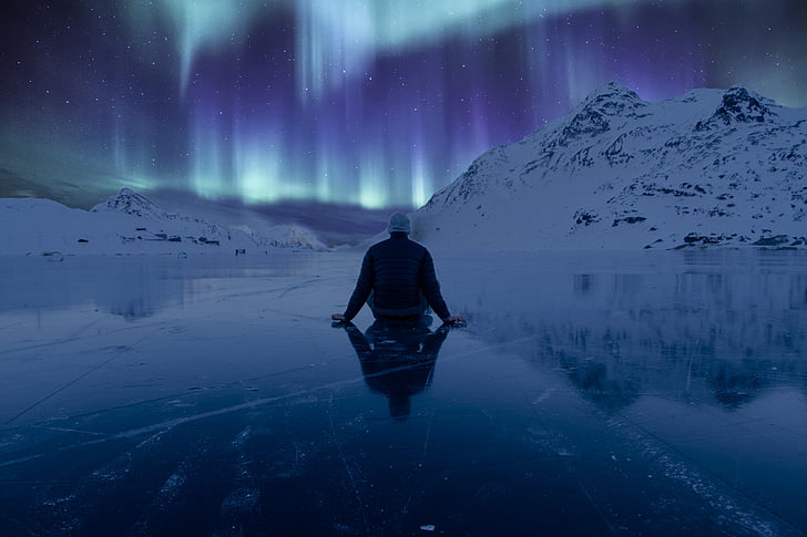 person sitting on ice surface watching aurora lights