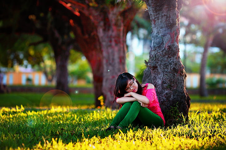 woman in pink cold-shoulder and green pants sitting on grass under brown tree during daytime