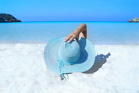 person holding blue wicker sun hat while laying on white sand during daytime