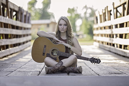 selective focus photography of woman wearing sleeveless top holding cutaway acoustic guitar sitting on bridge