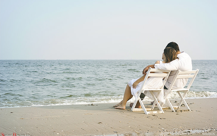 man and woman sitting on white wooden chairs beside the shore