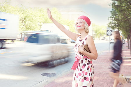 woman wearing white, pink, and brown polka-dot skater dress and red cap standing beside street waving her hand