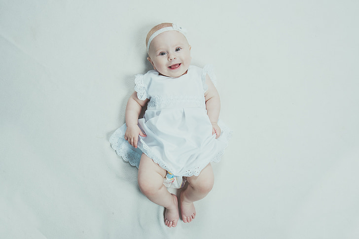 baby smiling wearing white headband and dress lying on bed