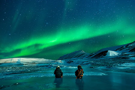 two people partially submerged on water surrounded by snow overlooking aurora borealis