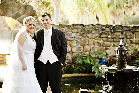 man in black suit and woman in white wedding dress standing near garden fountain during daytime