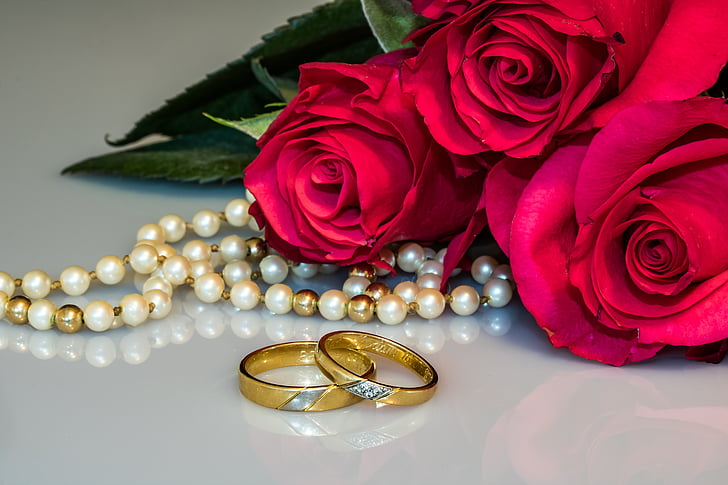 white and gold pearl necklace beside two gold-colored wedding bands and pink roses