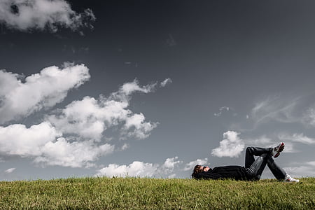 man lying on green grass field under cloudy sky