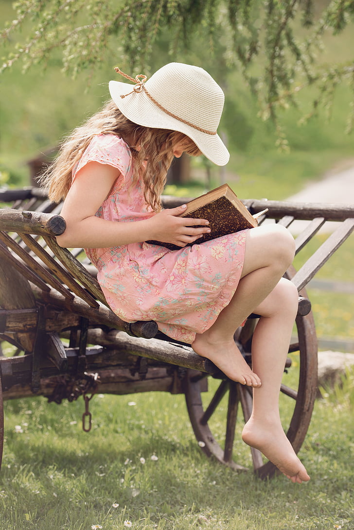 girl in pink and multicolored floral dress holding book sitting on brown wooden carriage