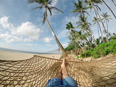 person with blue shorts lying on brown mesh hammock on seashore with blue sky and white clouds during daytime close-up photography