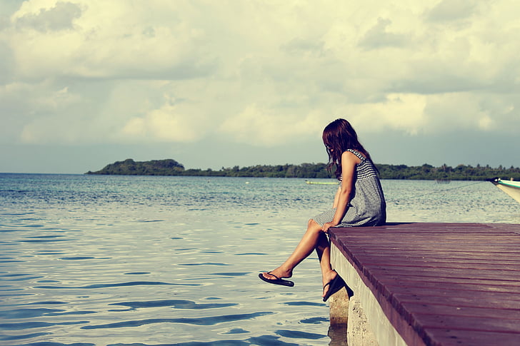 woman in gray dress sitting on dock during cloudy daytime
