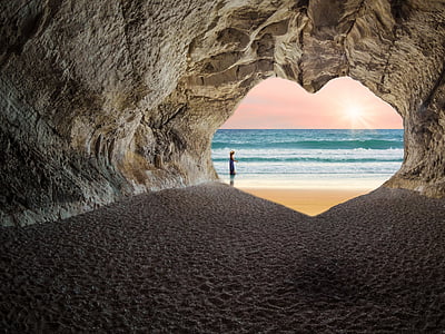 person standing outside heart-shaped cave entrance near sea