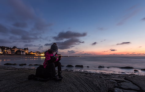 woman in pink and black winter suit sitting near body of water during yellow sunset
