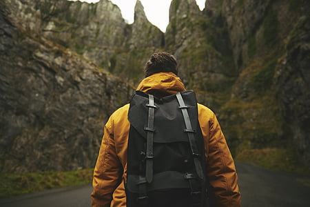 man wearing yellow hoodie and backpack standing in-front of a rocky mountain