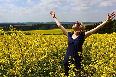 woman in blue scoop-neck dress smiling while looking in sky besides yellow petaled flower field during daytime