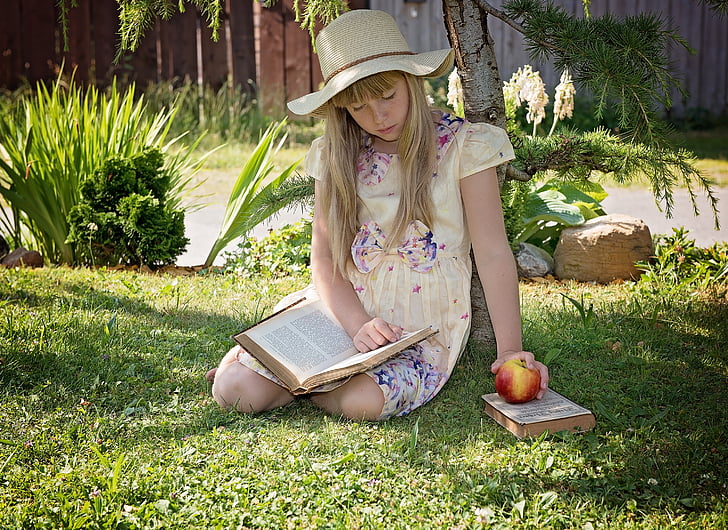 girl wearing dress sitting on grass holding apple and reading book