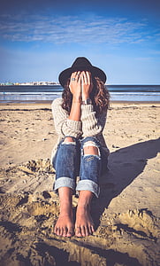 woman in gray sweater and blue denim distressed pants covering face with hands sitting on sand near body of water