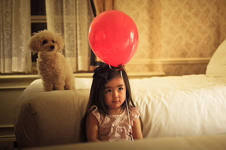 girl with red balloon beside a white poodle on bedroom