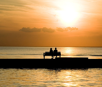 silhouette of two person sitting on bench near body of water during daytime