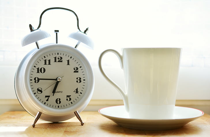 closeup photo of white ceramic mug on saucer beside white twin-bell alarm desk clock on brown wooden table