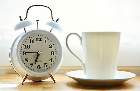 closeup photo of white ceramic mug on saucer beside white twin-bell alarm desk clock on brown wooden table