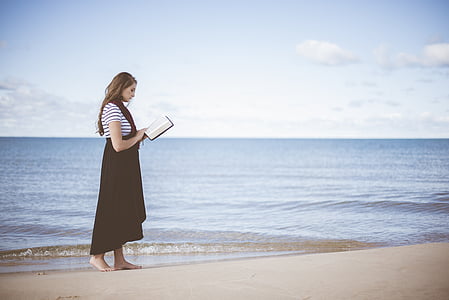 woman holding book while standing at seashore during daytime