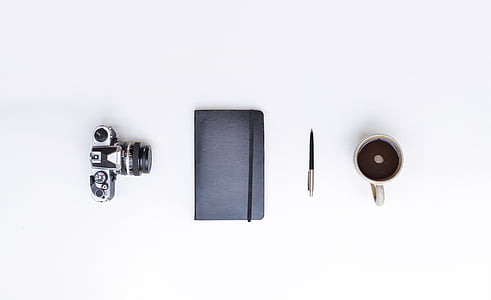 photo of DSLR camera, black handbook, pen, and mug filled with coffee on white surface