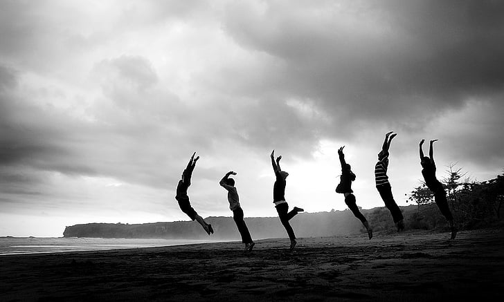 grayscale photography of six people jumping at sand nearby body of water