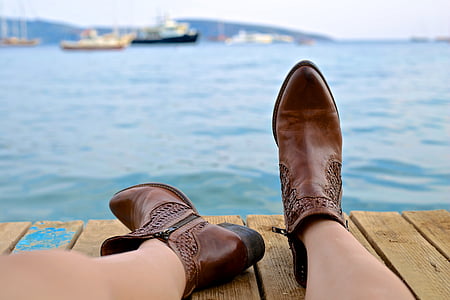 woman wearing brown leather boots near body of water and boats at daytime
