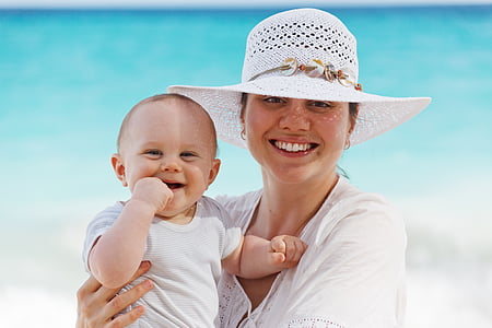 woman with white top and sun hat holding baby wearing white crew-neck shirt near shore