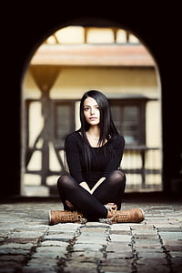 woman wearing black crew-neck top ad leggings and brown leather boots sitting on grey brick floor