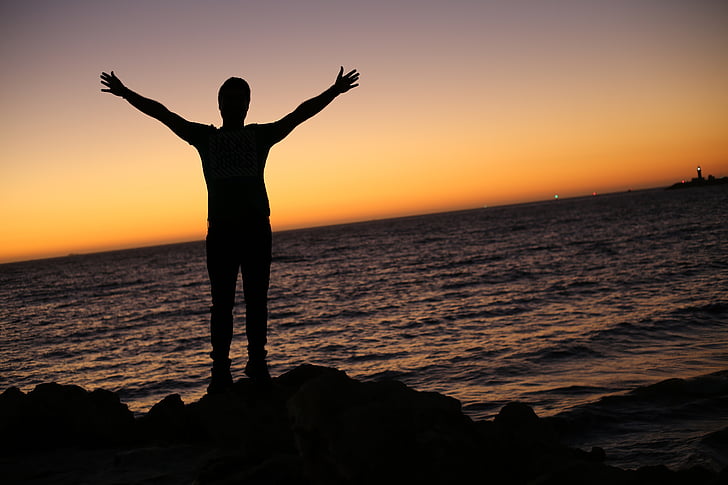 silhouette photo of person beside seashore raising hand during sunset
